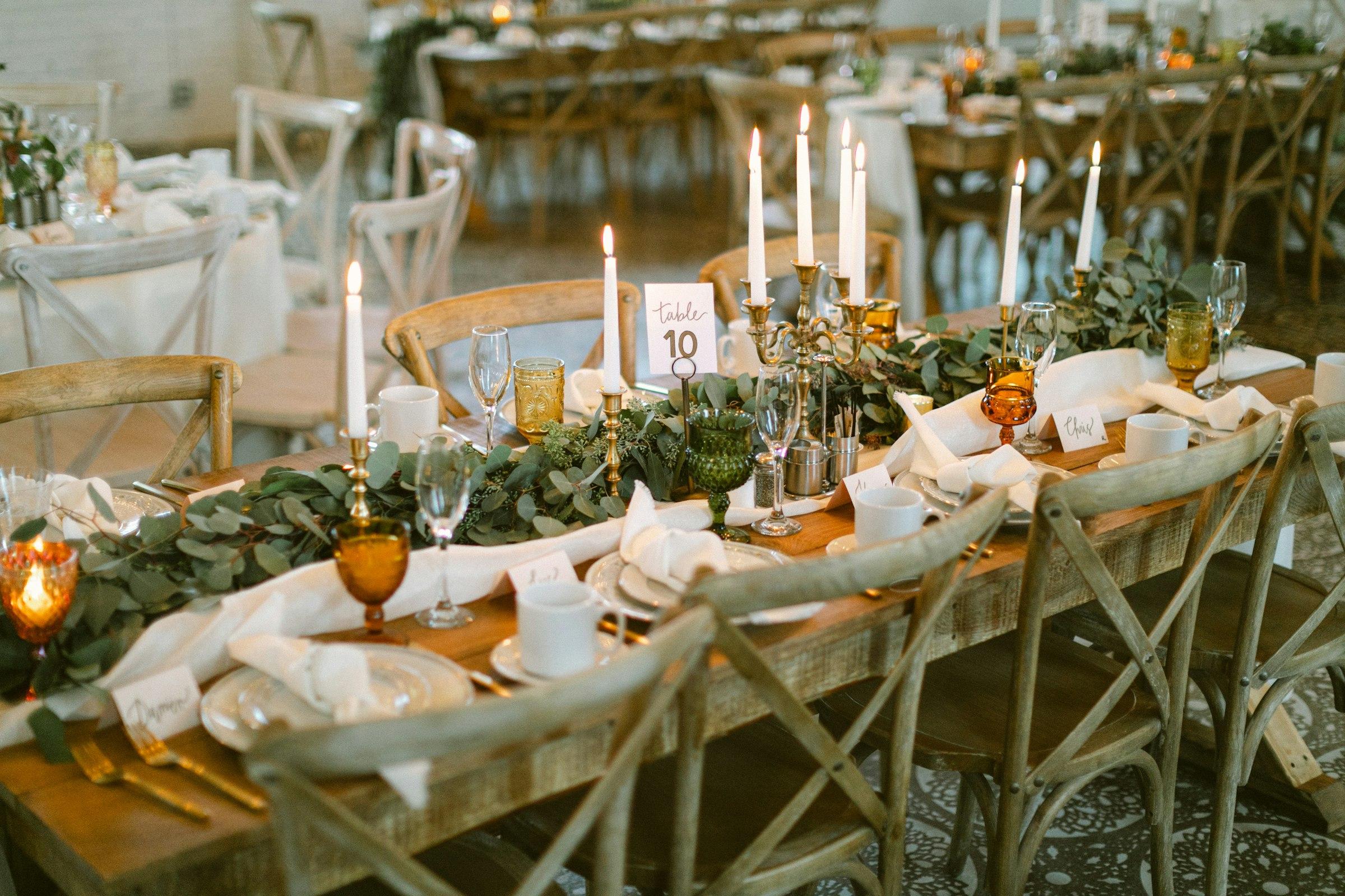 Longue table de réception dressée aux bougies, guirlande d’eucalyptus et chaises en bois